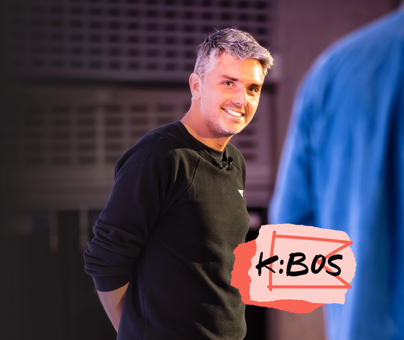 A person smiling, wearing a black shirt with a microphone attached. The text "K:BOS" is displayed in the foreground.