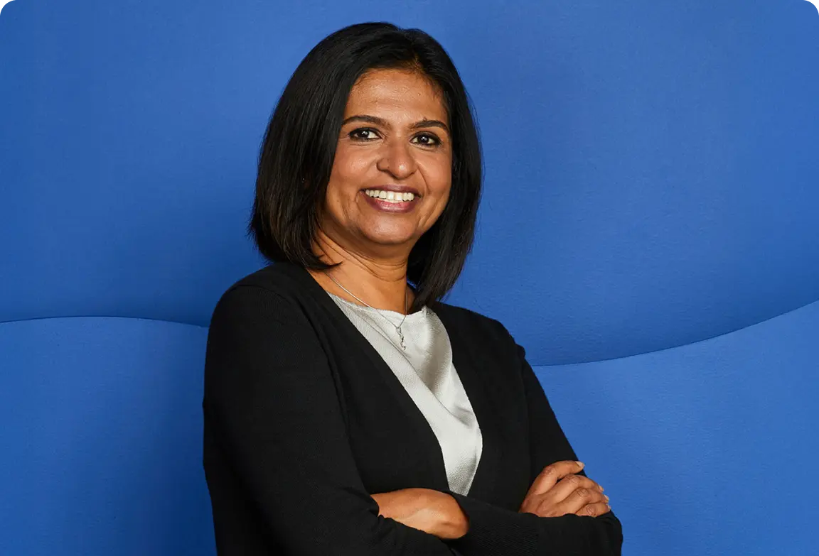 Smiling woman with shoulder-length hair, wearing a black cardigan over a light blouse, standing against a blue background.